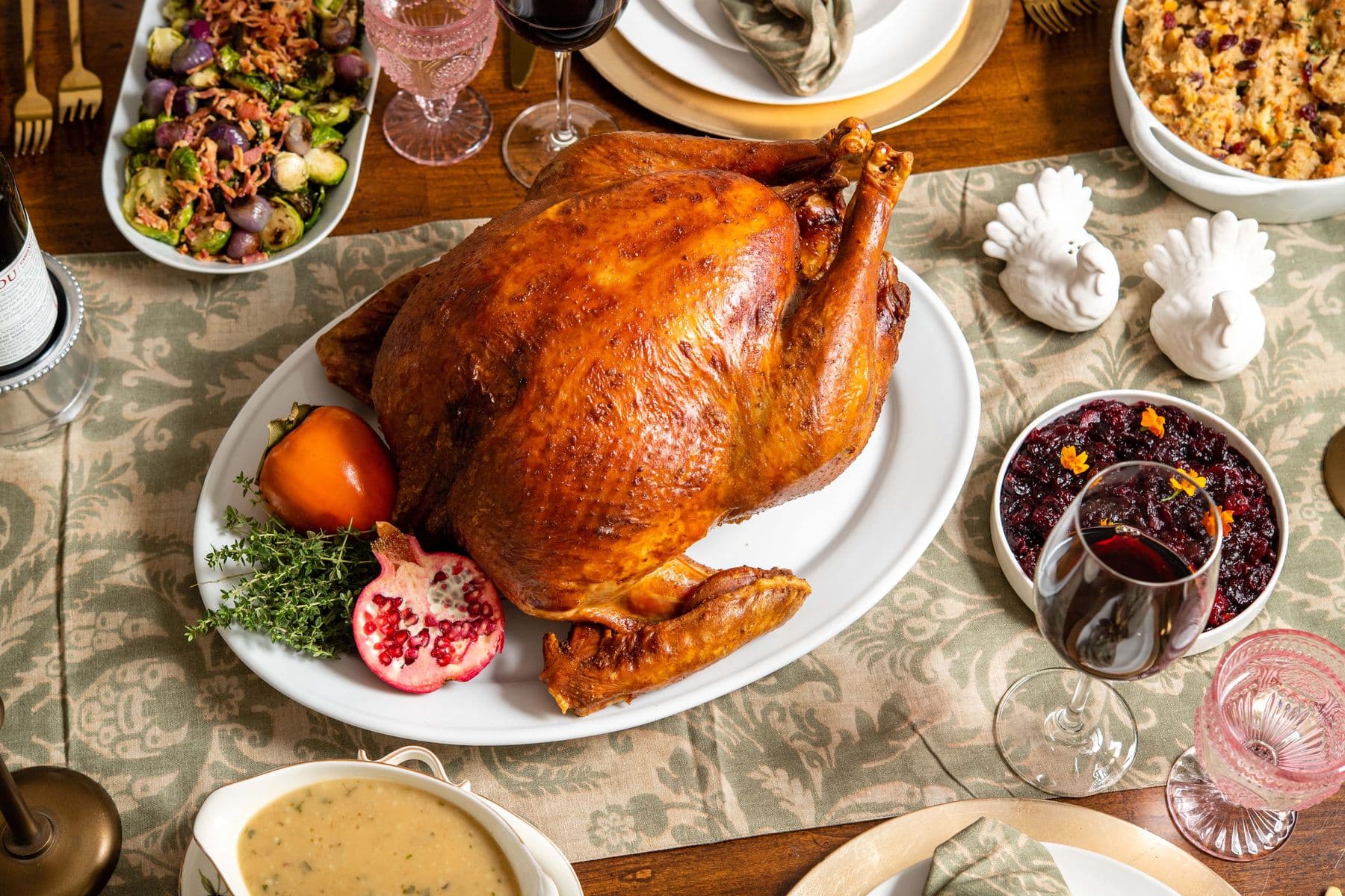 A whole herb-roasted turkey on a platter, surrounded by Brussels sprouts, cranberry sauce, gravy and stuffing on a set holiday table